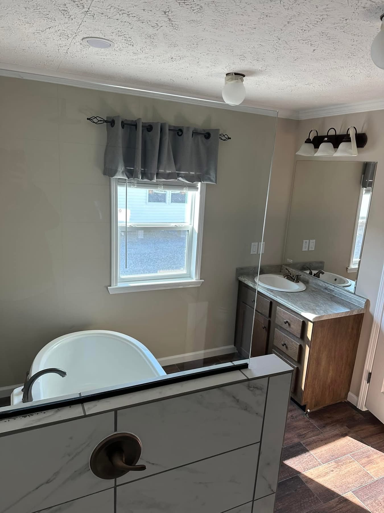 Modern bathroom with a white bathtub behind a marble wall. Dark wood vanity with a dual sink and large mirror. Window with gray curtain. Warm, inviting ambiance.