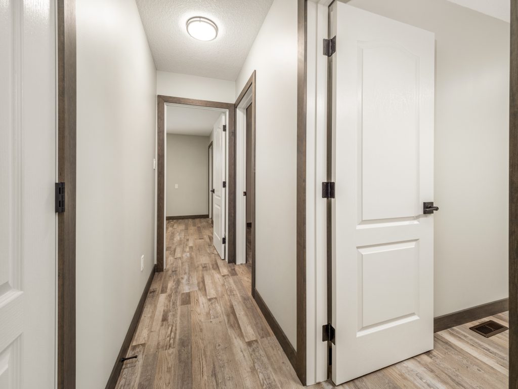 A well-lit hallway with light wood flooring and beige walls. Three white doors with dark trim are open, leading to other rooms. A ceiling light provides illumination.
