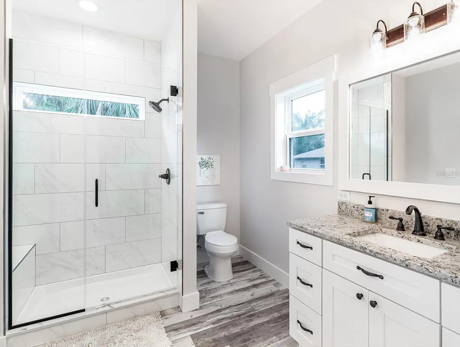 Bright bathroom with a glass-enclosed shower, white tiles, a toilet, and a granite countertop with dual sinks. A window and light fixtures add a fresh feel.
