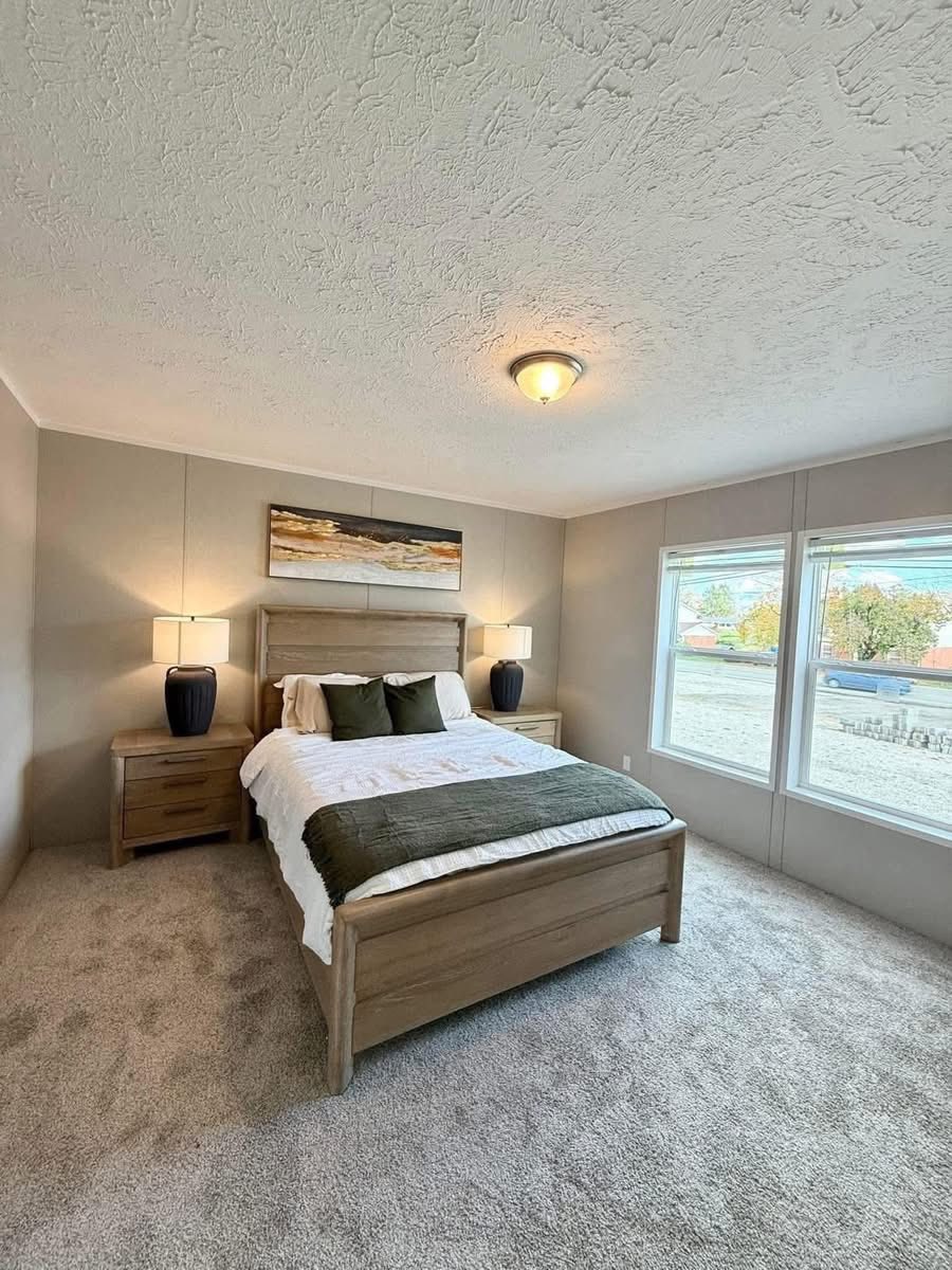 A cozy bedroom with a large bed, two bedside tables, and lamps. Neutral tones, soft lighting, abstract artwork, and windows create a serene ambiance.