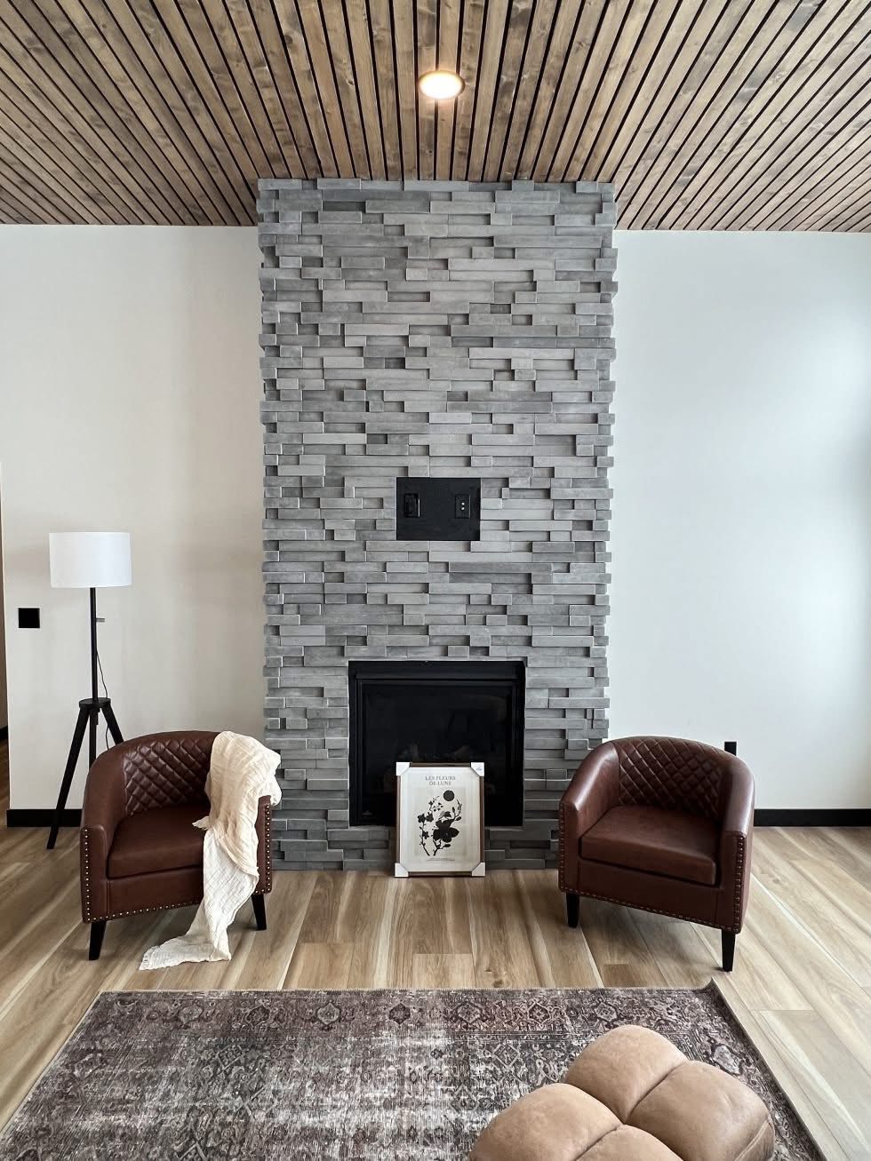 Modern living room with a gray stone fireplace, two brown leather chairs, and a floor lamp. A beige rug and plush ottoman add warmth. Cozy ambiance.