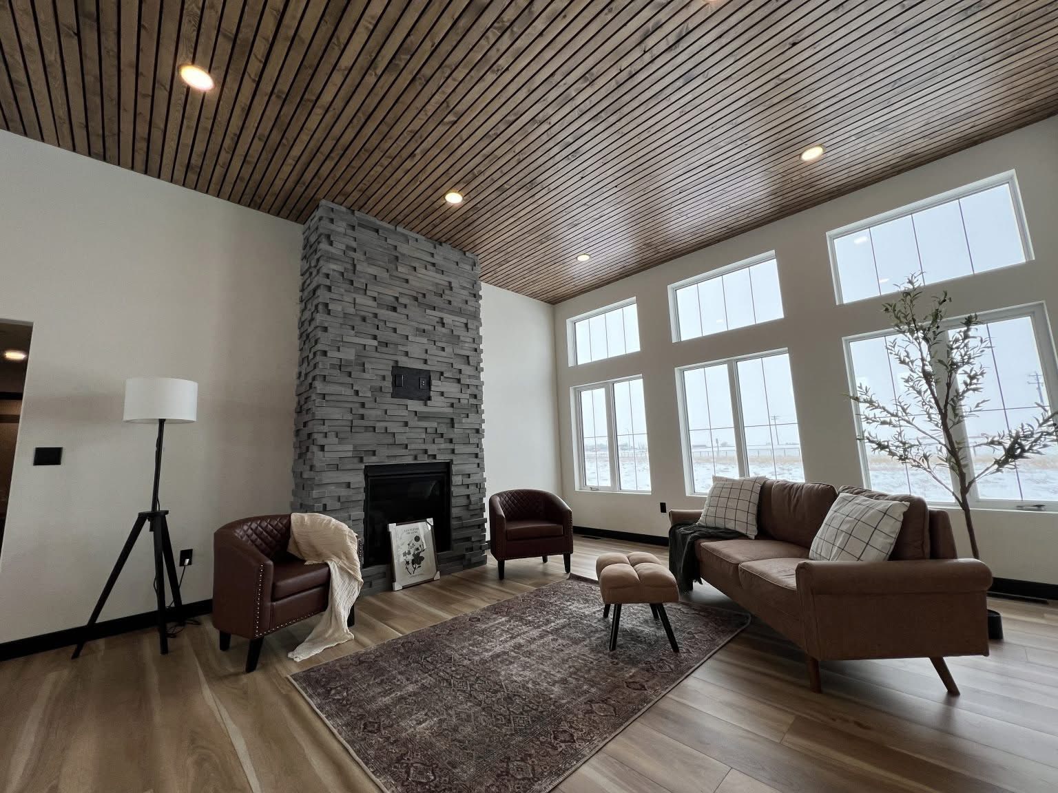 Modern living room with a cozy feel, featuring a stone fireplace, brown leather furniture, large windows, wooden ceiling, and stylish decor.