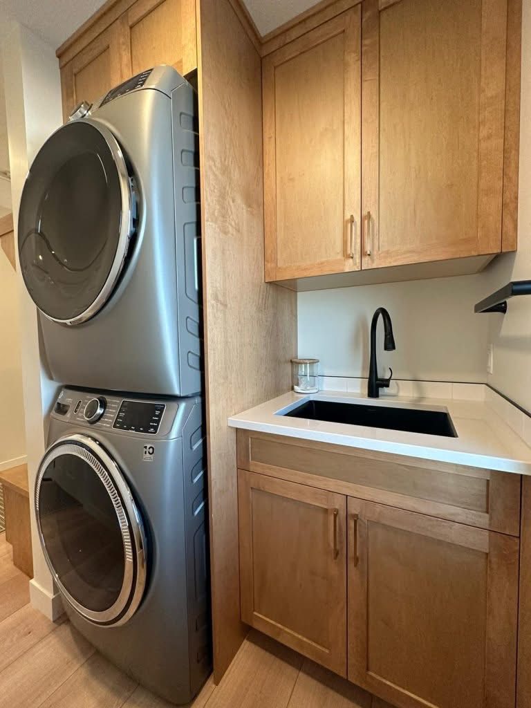 A compact laundry room with a stacked silver washer and dryer on the left. To the right, light wood cabinets and a sleek black faucet over a white sink. Calm and efficient.
