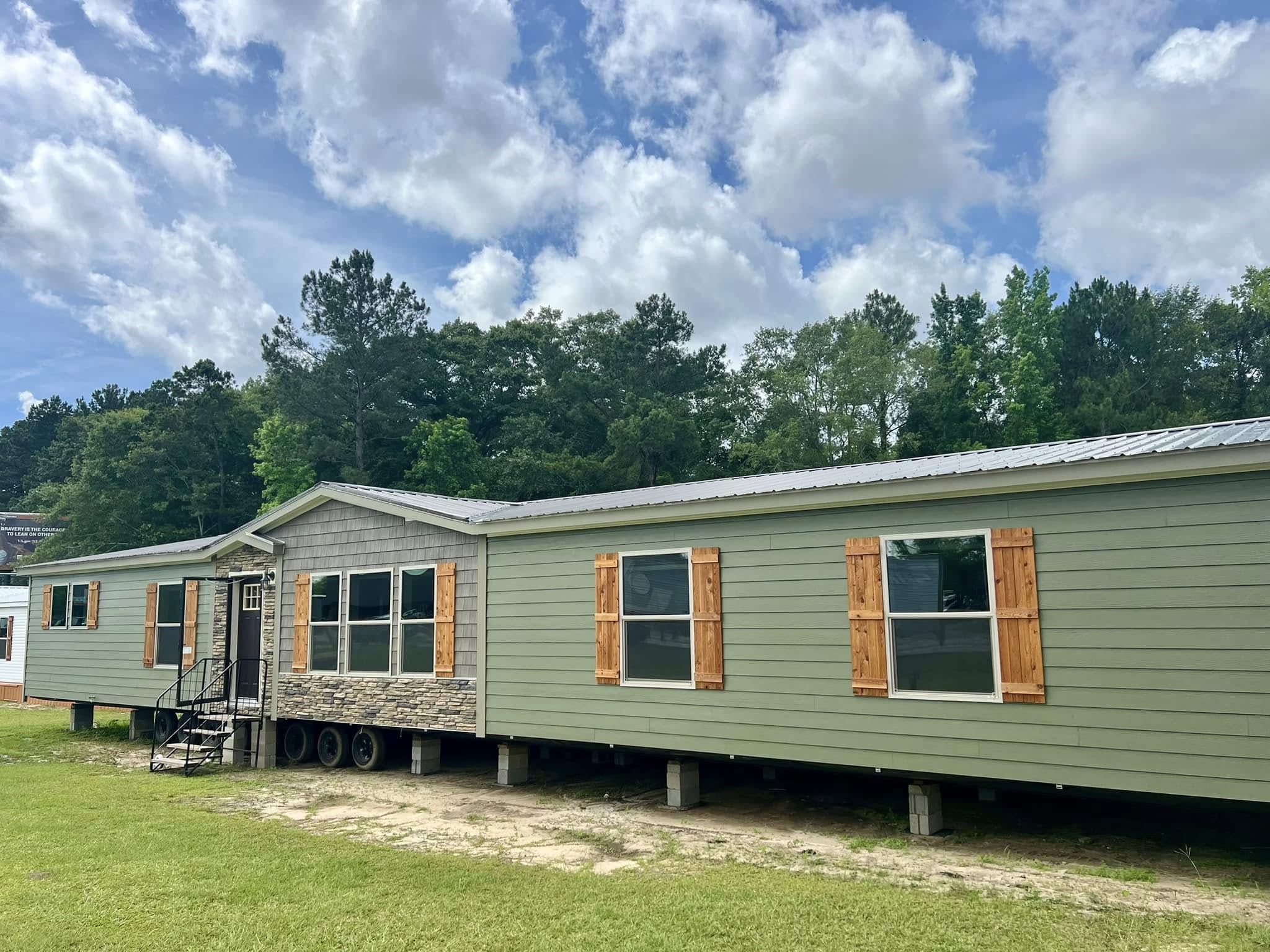 A green mobile home with wooden shutters and stone accents sits on a grassy area under a blue sky with scattered clouds, surrounded by lush trees.