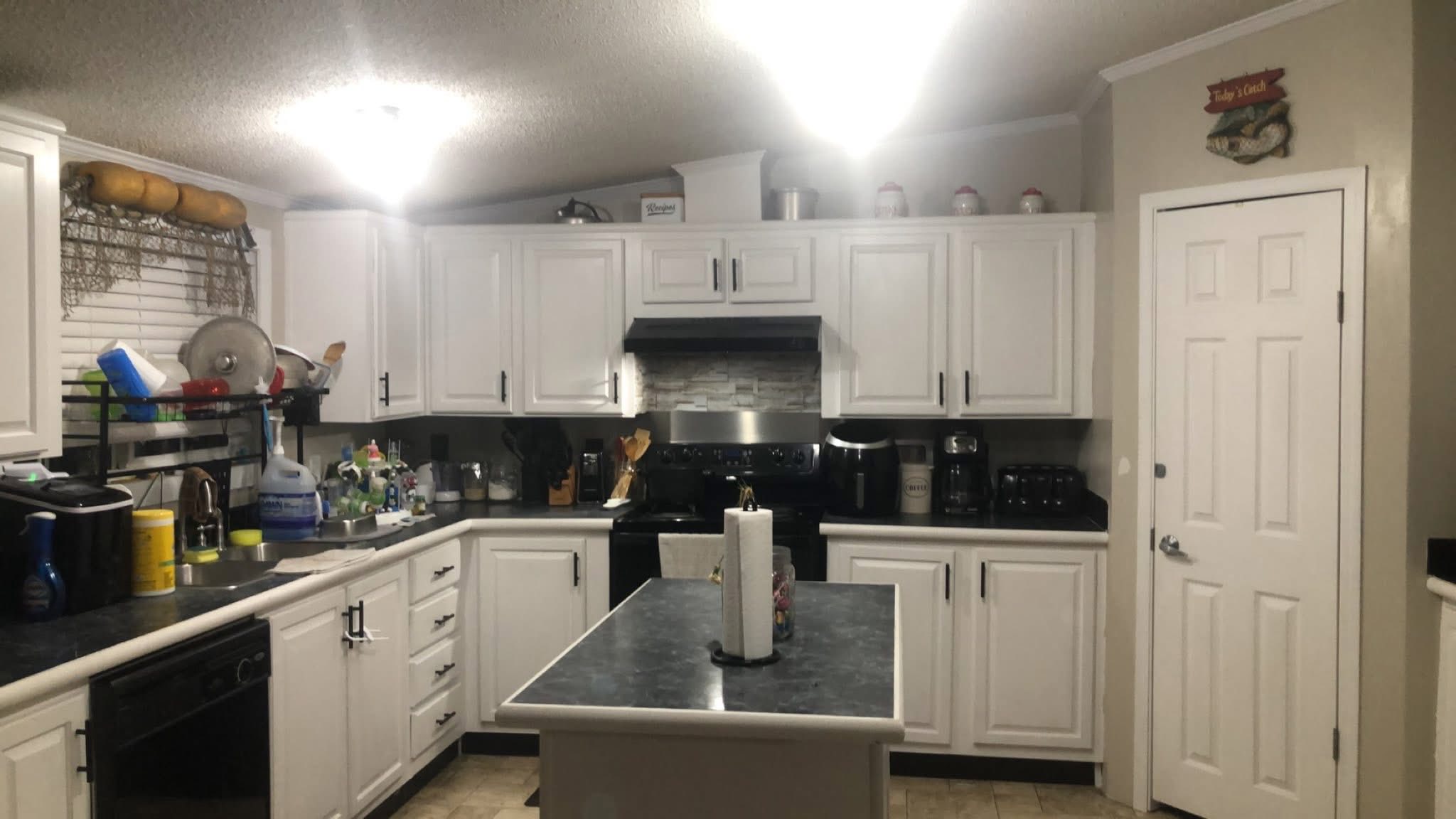 A well-lit kitchen with white cabinets and black countertops. Central island with paper towels, appliances on counters, organized, tidy, and inviting.