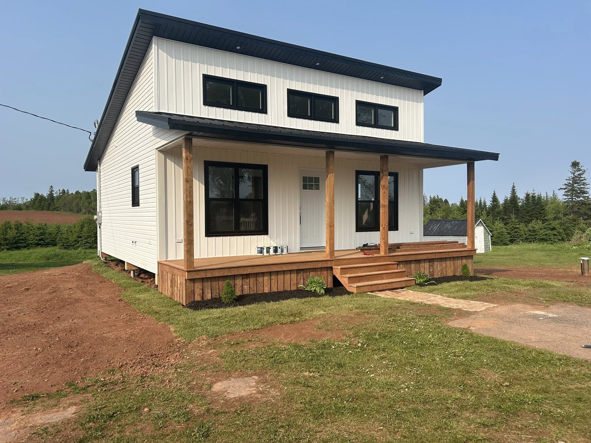Modern white house with a black roof and wooden porch in a rural setting. Large windows, neatly cut grass, trees, and clear sky create a serene atmosphere.