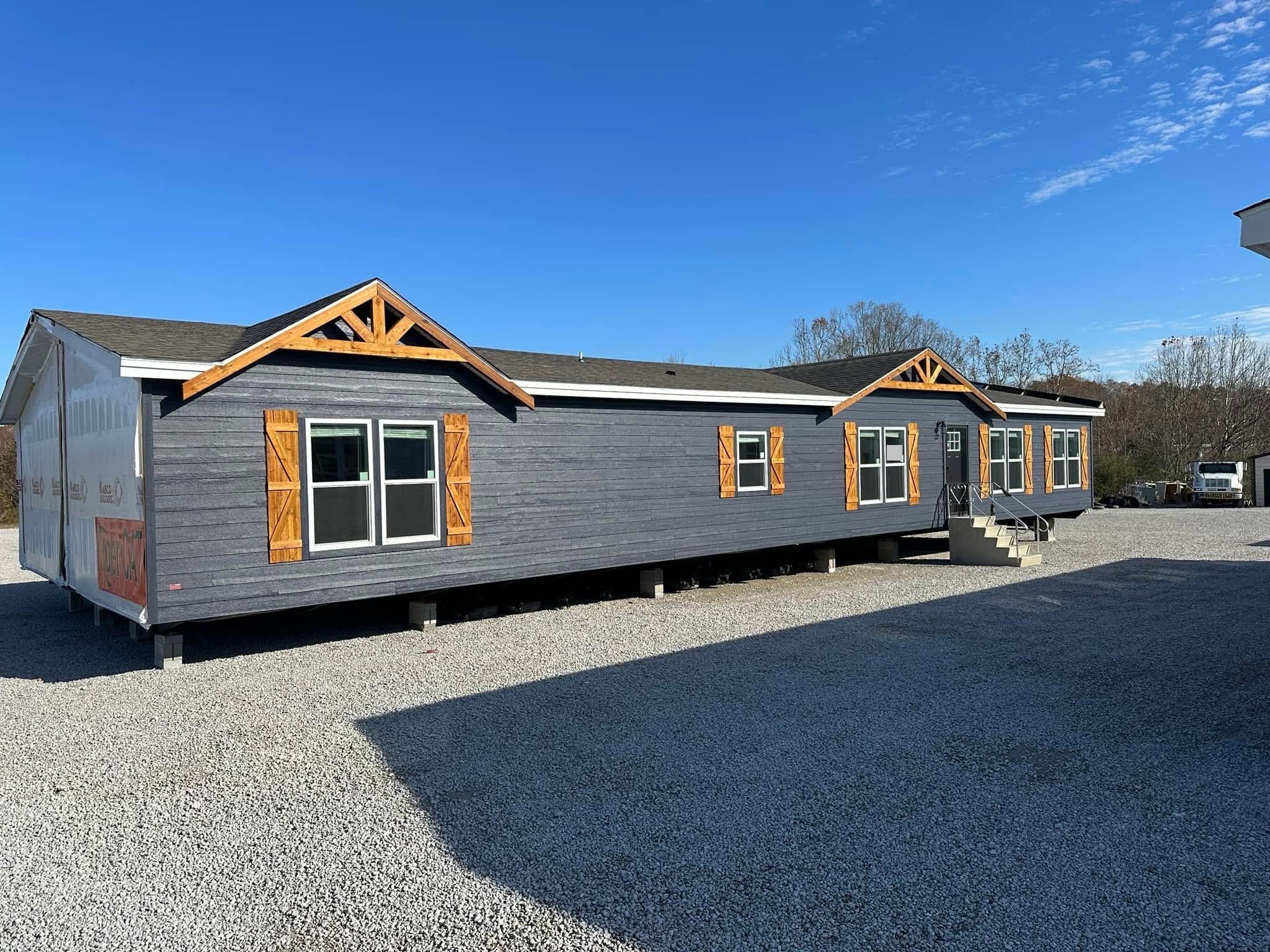 A long, gray modular home with wooden trim and shutters sits on a gravel lot under a clear blue sky. Steps lead to the entrance, conveying a calm, sunny atmosphere.