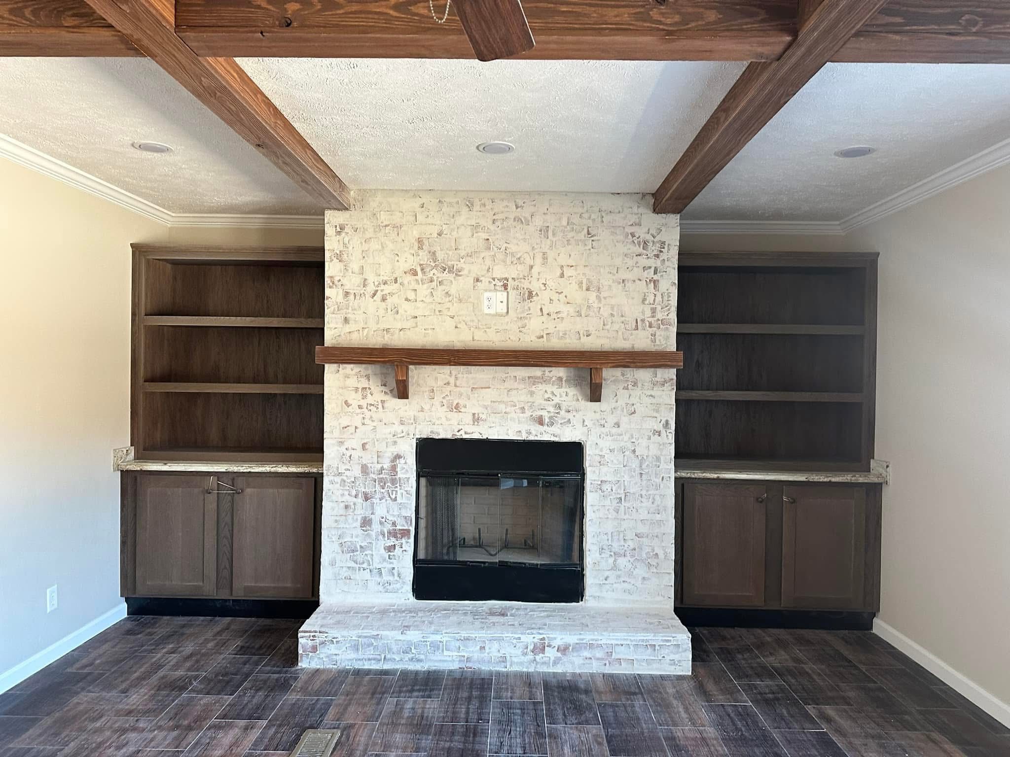 A room features a whitewashed brick fireplace with a wooden mantel, flanked by dark wood cabinets and bookshelves, set against a ceiling with wooden beams.