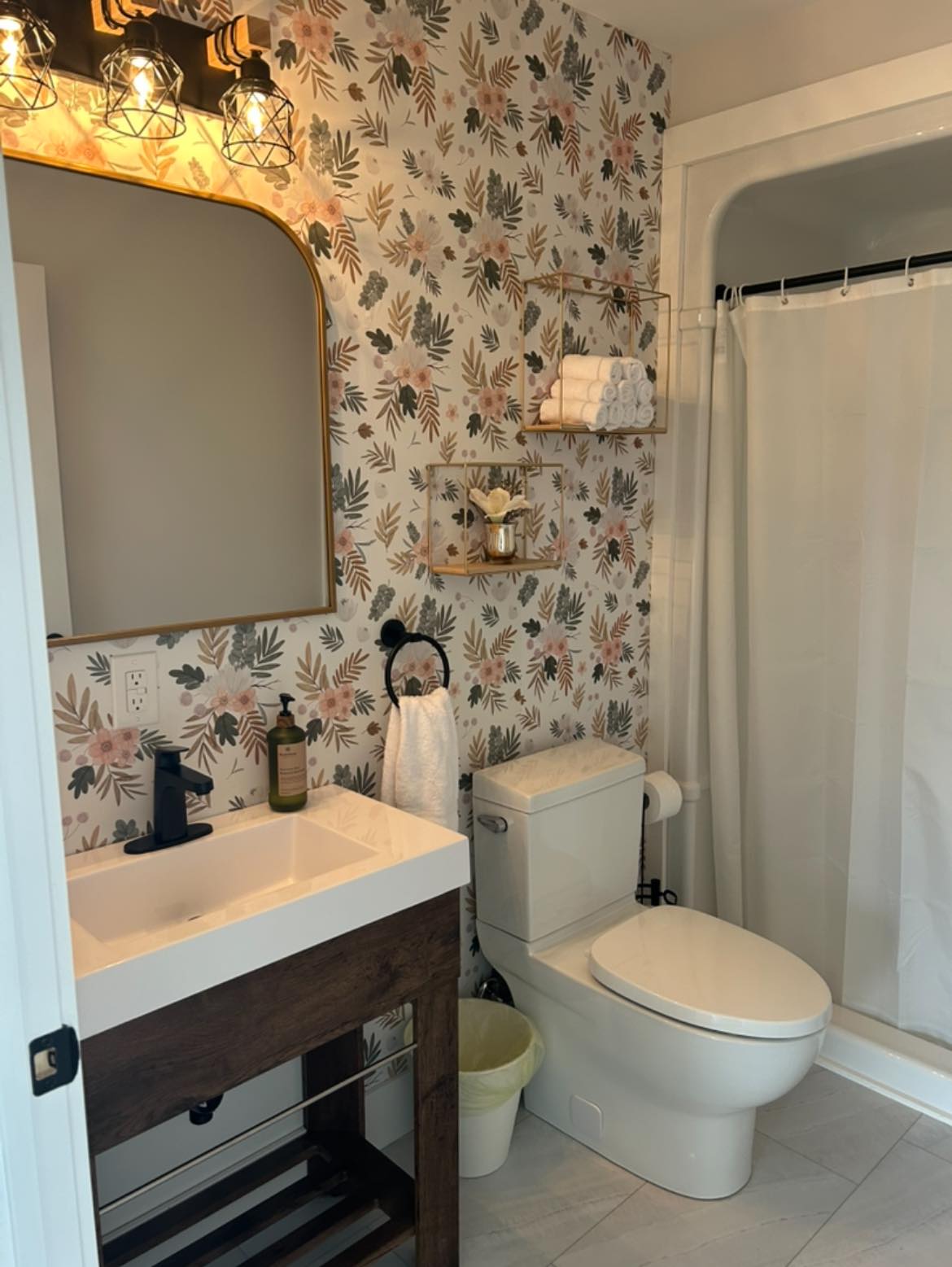 A cozy bathroom with floral wallpaper, a wooden vanity with a square sink, and industrial light fixtures. Shelves hold rolled towels and decor. The atmosphere is warm and inviting.