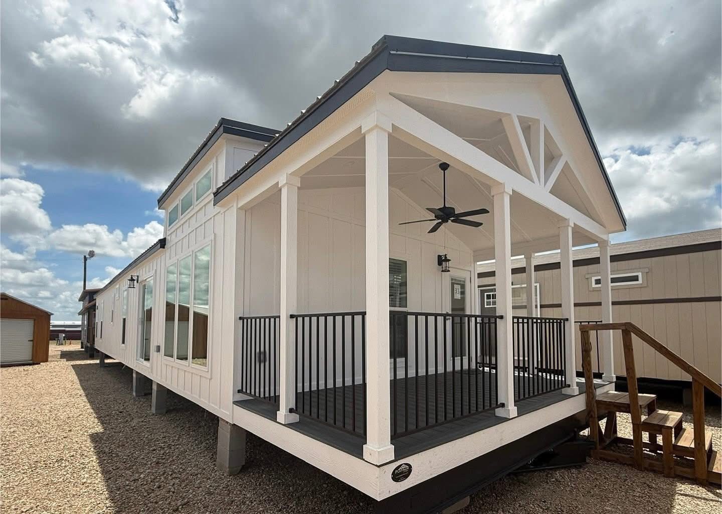 Modern tiny house exterior with a white facade and large windows. Features a covered porch with black railing and ceiling fan, under a partly cloudy sky.