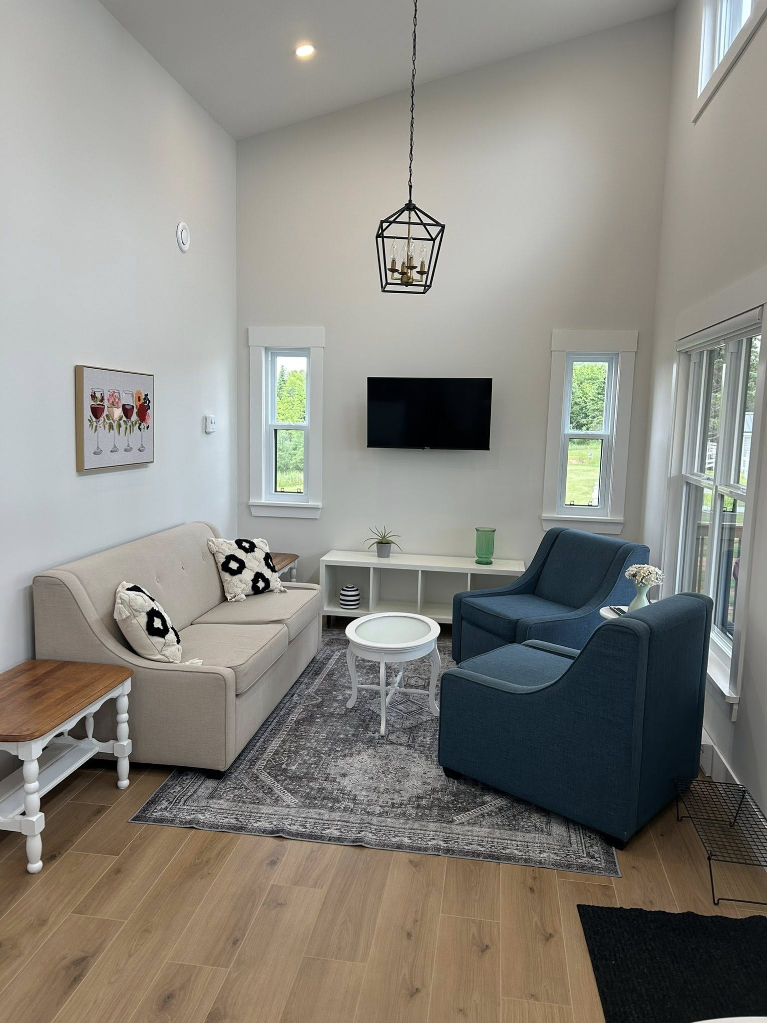 A modern living room with high ceilings, featuring a beige sofa with patterned pillows, two blue armchairs, and a small round white table on a gray rug. A flat-screen TV is mounted between two windows with green outdoor views. A geometric pendant light hangs from the ceiling, and a wooden side table holds a potted plant. The room exudes a cozy, minimalist feel.