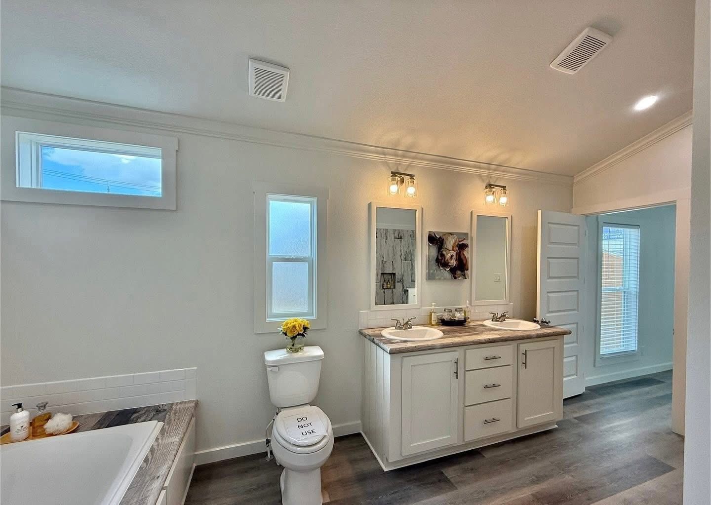 Spacious bathroom with double sinks, mirrors, and soft lighting. A bathtub is on the left, and a toilet with a "Do Not Use" sign is nearby. Bright and inviting.