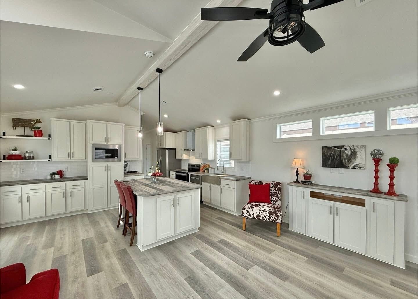 Modern kitchen interior with white cabinets, a central island with red chairs, light wood flooring, and ceiling fan. Bright and inviting atmosphere.