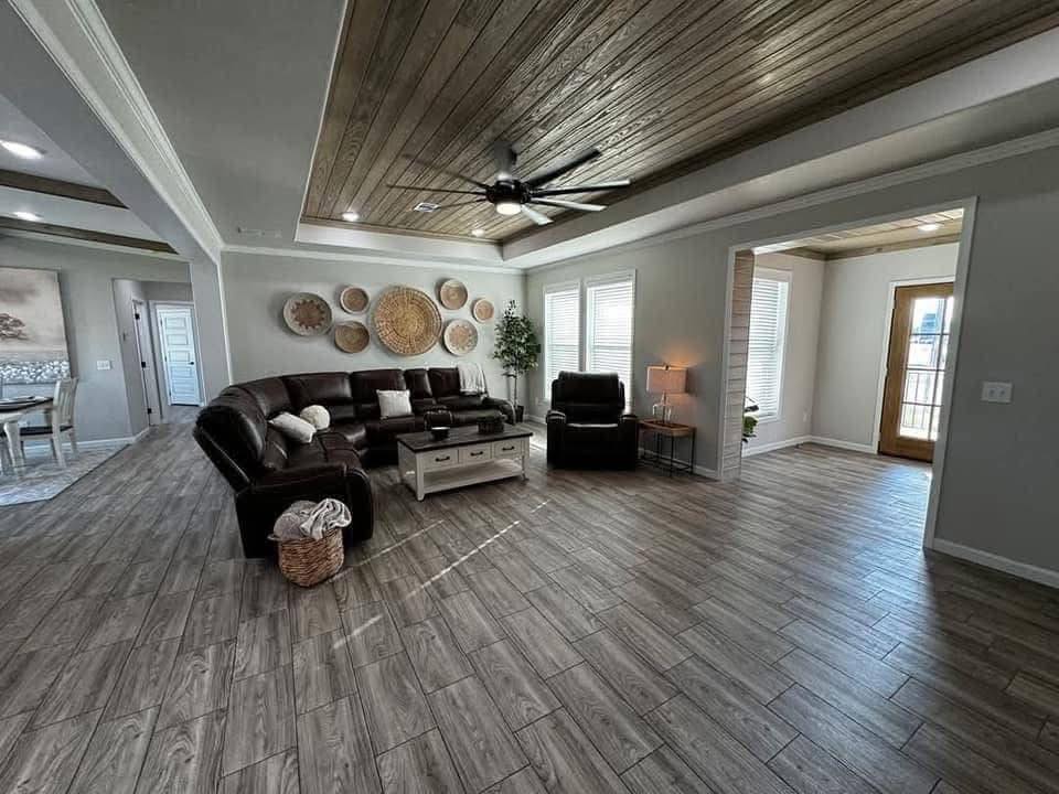 Spacious living room with brown leather sectional sofa, basket wall art, wooden ceiling, gray wood flooring, and natural light from large windows. Cozy and modern.