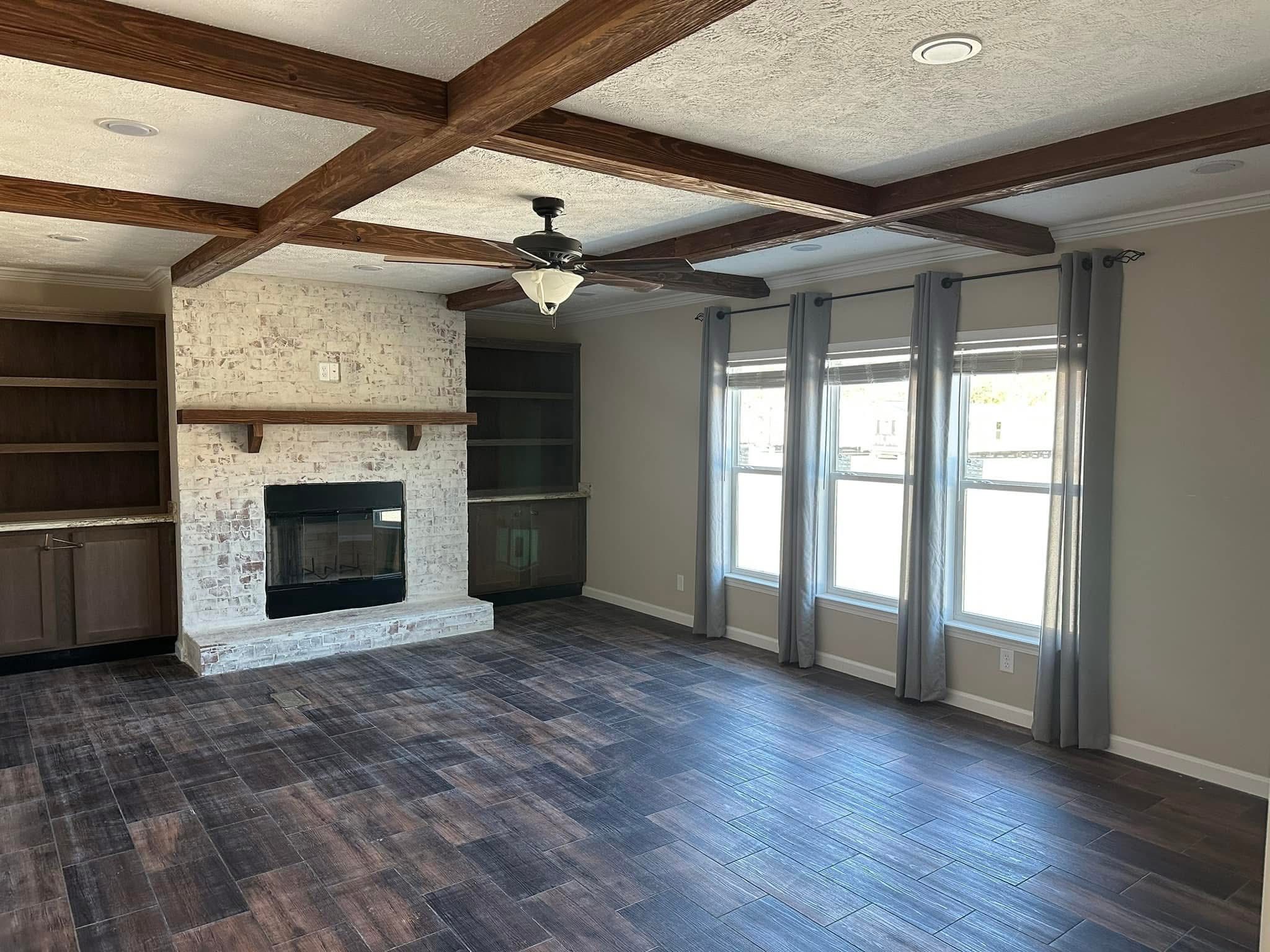 Spacious living room with wooden beams on the ceiling, a stone fireplace, and dark wood floors. Large windows with gray curtains add natural light.