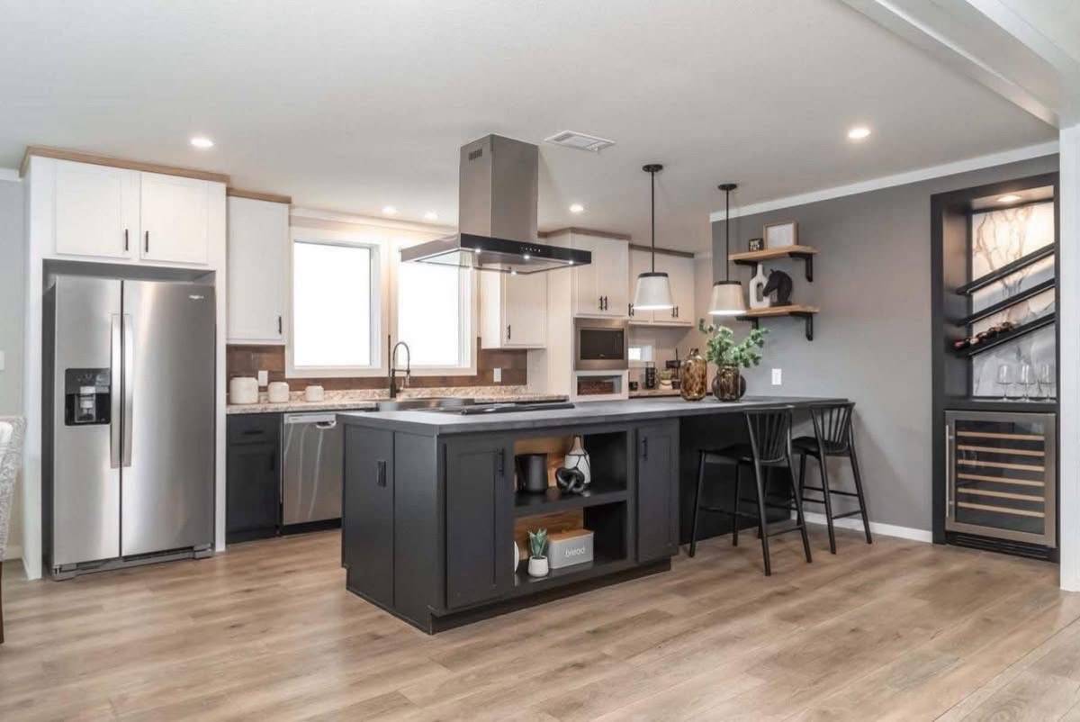 Modern kitchen with a central island, dark cabinetry, and a sleek range hood. Pendant lights hang above, while a wine rack and bar stools add elegance.