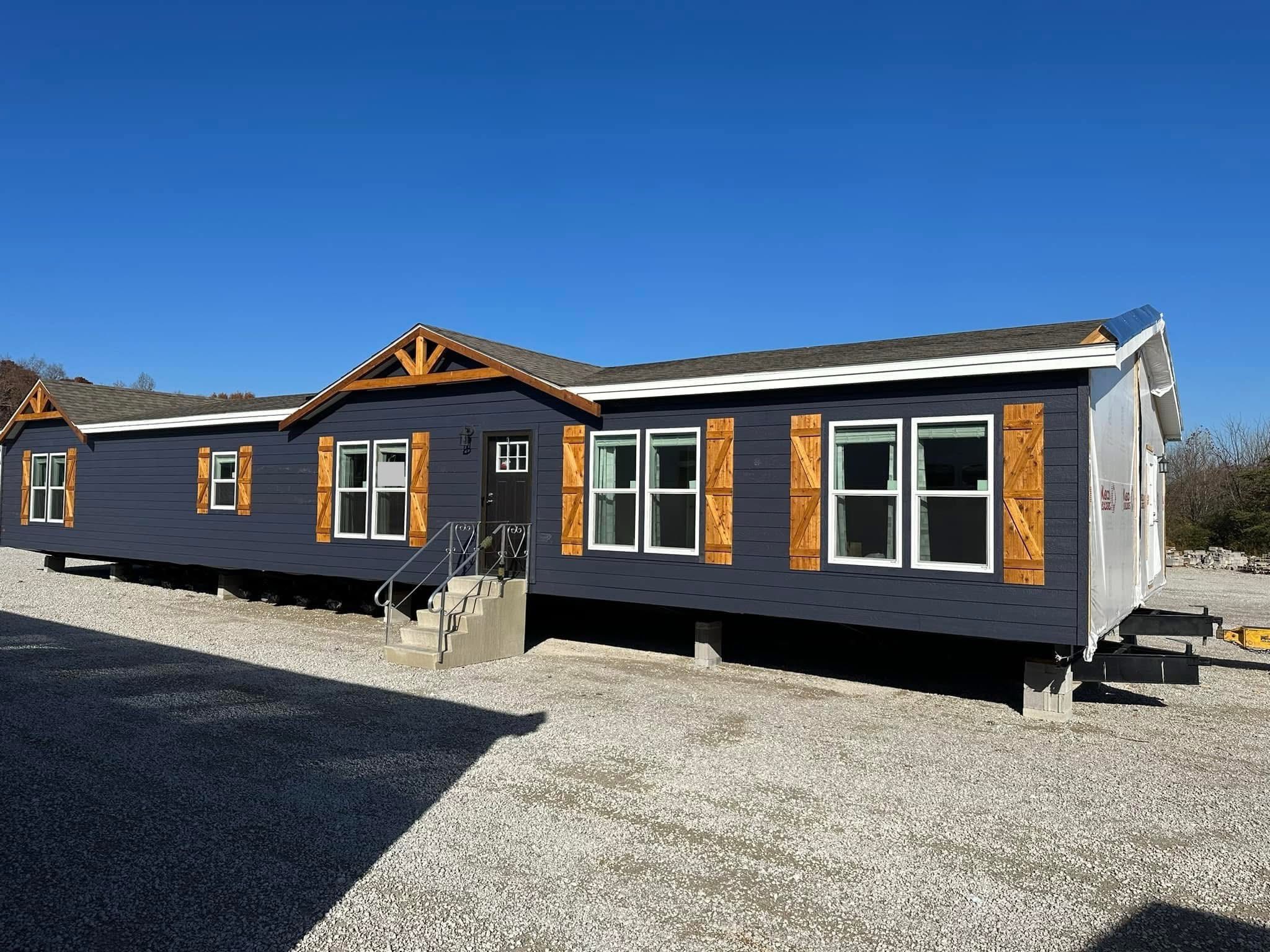 A modern, dark gray manufactured home with wooden shutters and decorative gables sits on a gravel lot under a clear blue sky, conveying a sense of simplicity and functionality.