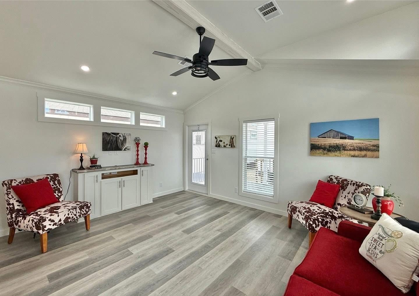 Bright living room with light wood flooring, white walls, and a vaulted ceiling. Red and floral armchairs, modern decor, and a ceiling fan create a cozy, inviting atmosphere.