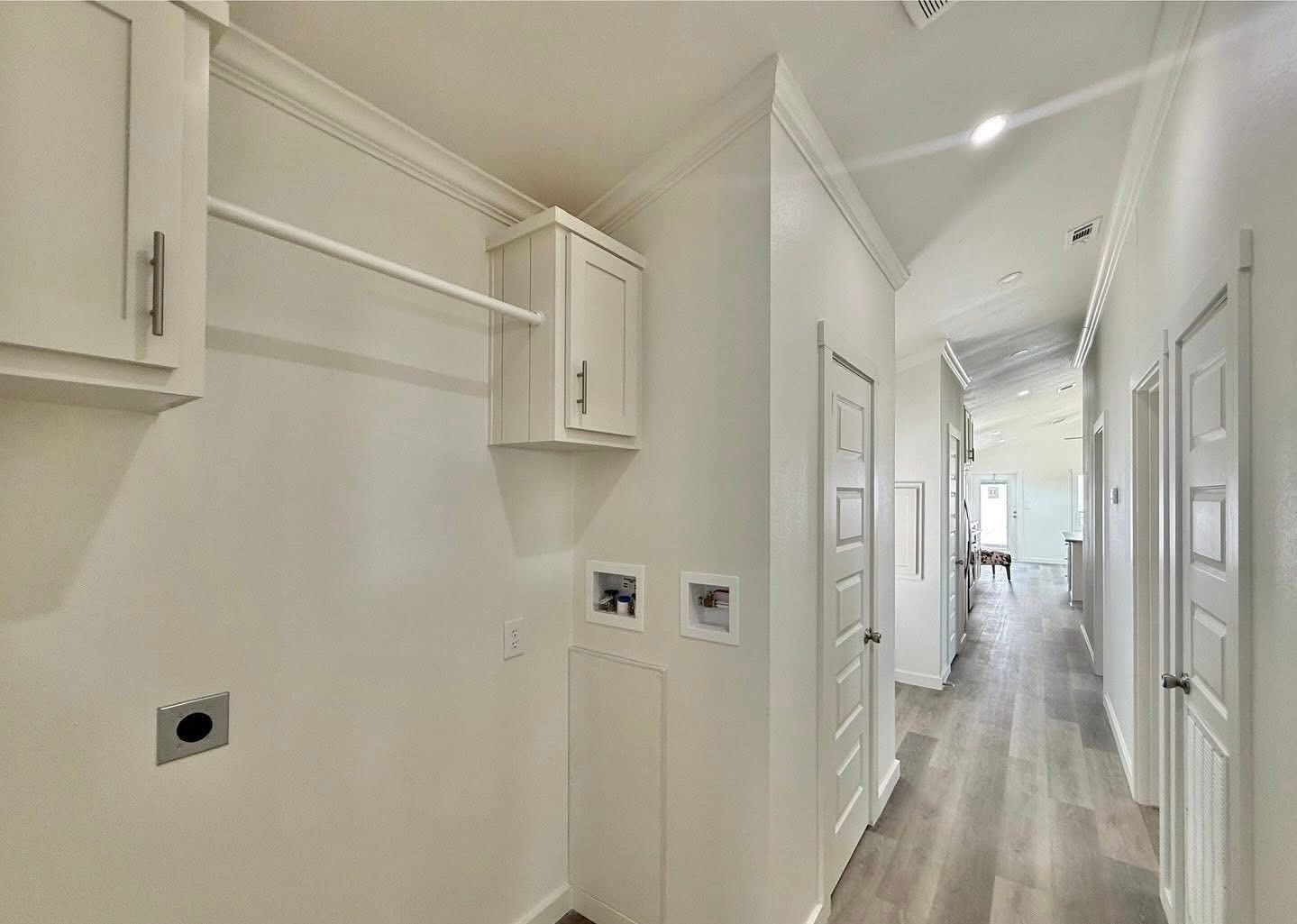Bright, modern hallway with white walls, recessed lighting, and wood flooring. Laundry area with cabinets on the left, doors leading to rooms on the right.