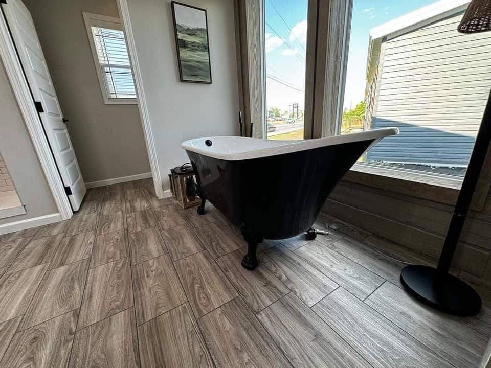 Freestanding black clawfoot bathtub with white interior, set on wood-patterned tiles near large windows. Soft natural light creates a serene, modern atmosphere.