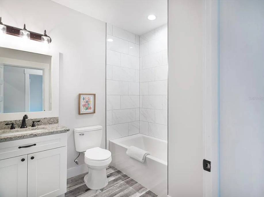 Modern bathroom with white tiles and a tub with a folded towel. A granite-topped vanity with a large mirror, light fixture, toilet, and wood floor create a clean, calm atmosphere.