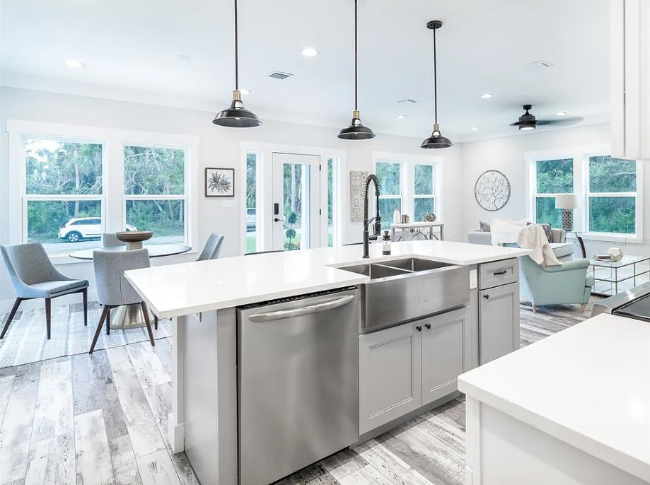 Modern open-concept kitchen with white cabinets, stainless steel appliances, and a large island. A cozy living area and dining space are visible, with natural light streaming in through large windows, creating an inviting atmosphere.