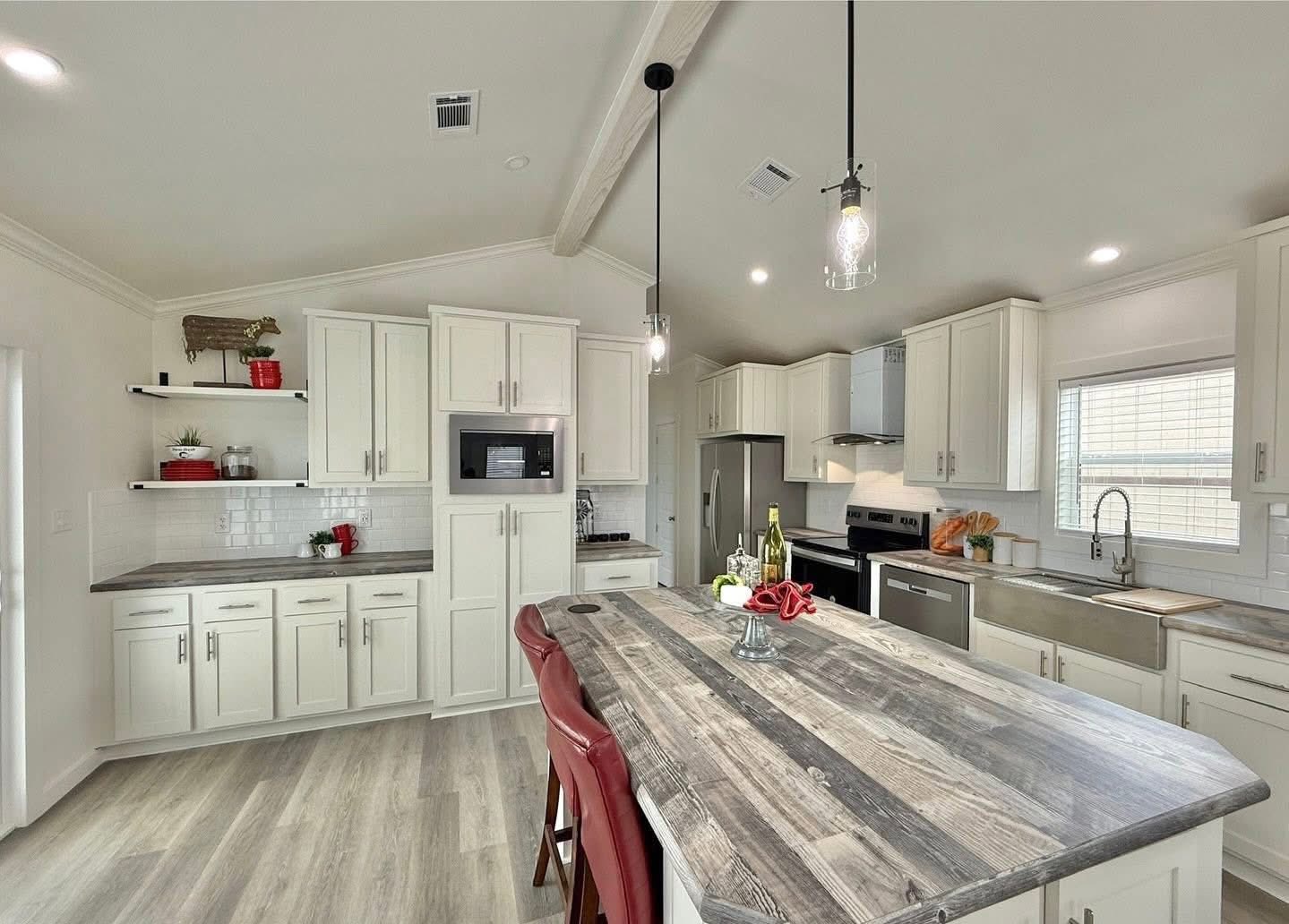 Modern kitchen with white cabinets, wood island, and red bar stools. Pendant lights create a bright, inviting atmosphere. Decor includes wine, glasses, and plants.