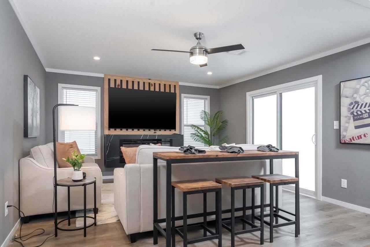 Modern living room with gray walls, featuring a beige sofa, wooden TV stand, large wall-mounted TV, and bar table with stools. Bright and cozy vibe.