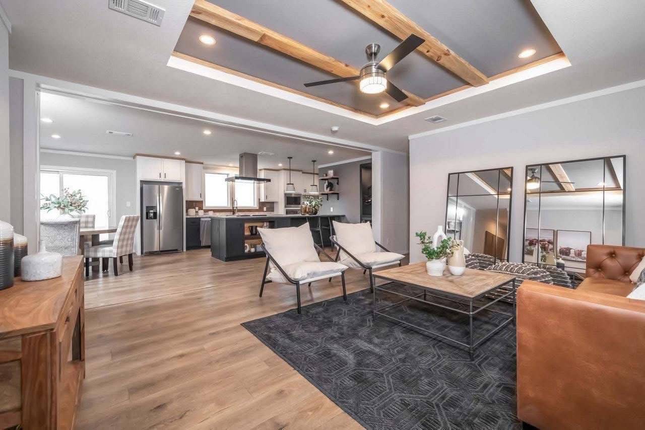 Modern living room with wood accents, featuring a brown leather sofa, glass coffee table, and mirrors. Kitchen and dining area visible in the background.