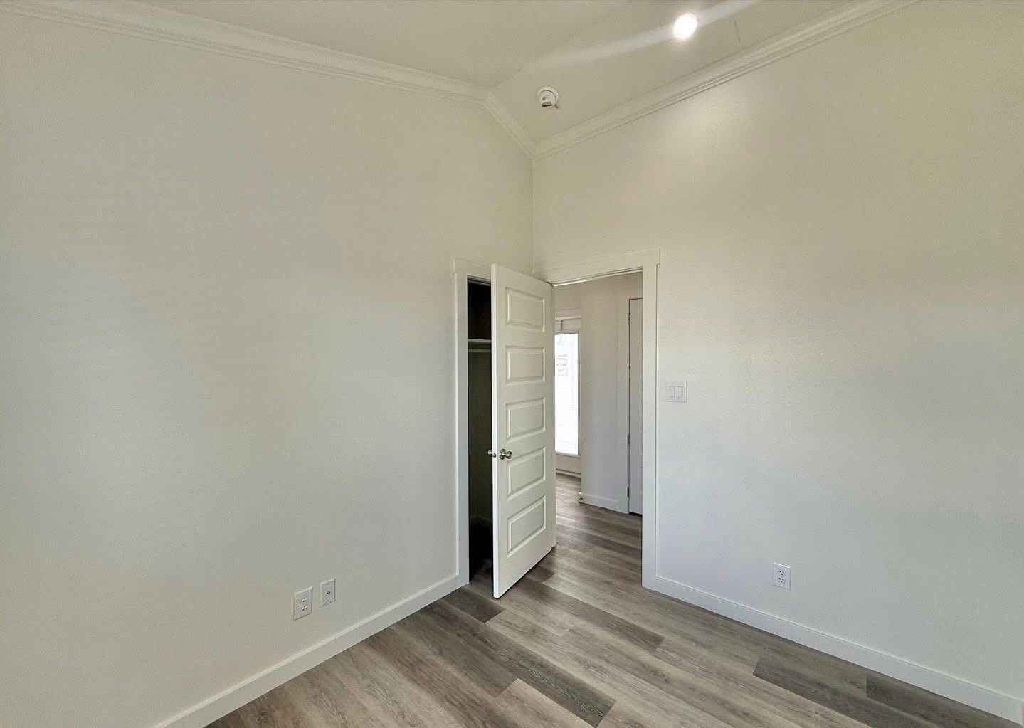 An empty room with light gray walls and dark wood flooring. An open door reveals a closet with a white frame. Soft light suggests a calm, welcoming ambiance.