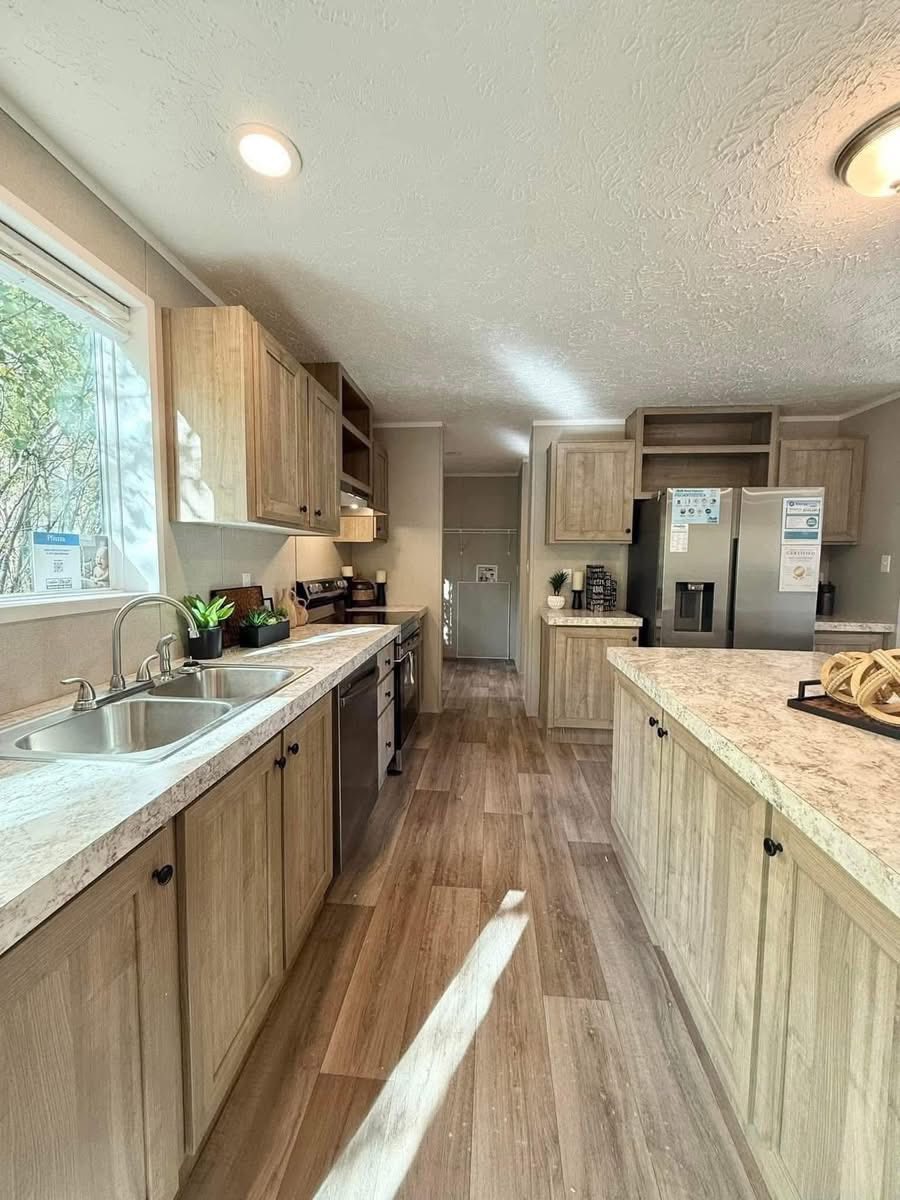 Spacious kitchen with light wood cabinets, stone countertops, and stainless steel appliances. Sunlight streams through a window, creating a warm atmosphere.