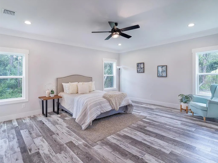 Spacious bedroom with gray and white decor, featuring a cozy bed, light wood floors, a ceiling fan, large windows, a pale blue chair, and a leafy plant.