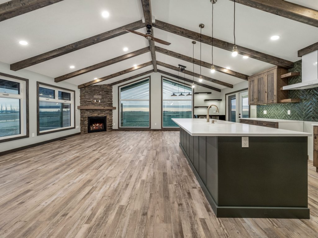 Spacious modern kitchen with vaulted ceiling, exposed beams, and large windows. Features a stone fireplace, wood flooring, and a central island, exuding warmth.