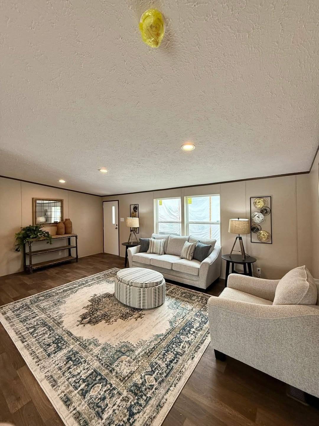 Cozy living room with a large beige and blue patterned rug, two sofas, a round ottoman, and warm lighting. Decor includes plants and wall art.