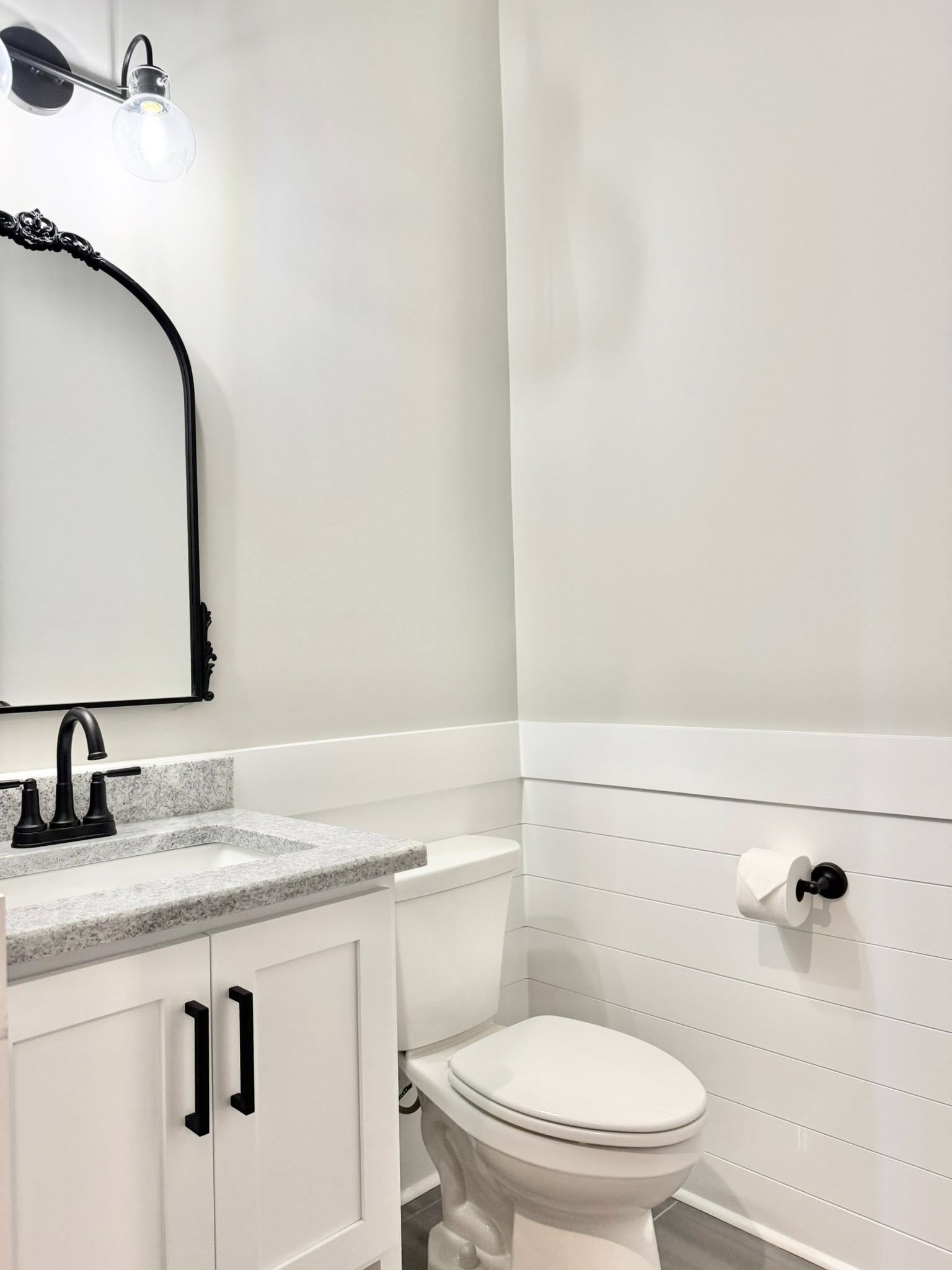 Small, modern bathroom with a white vanity, granite countertop, black faucet, and ornate mirror. Soft lighting creates a clean, minimalist feel.