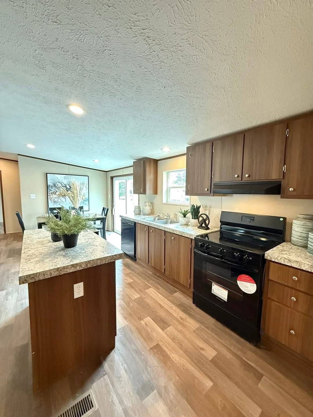 A modern kitchen features wood cabinets, black appliances, and a granite island with plants. Sunlight brightens the space, offering a welcoming feel.