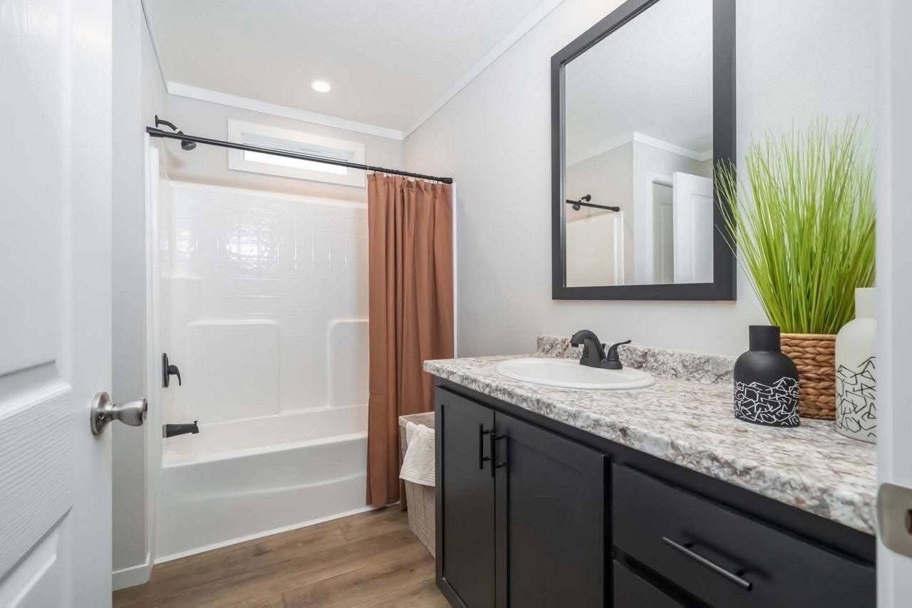 Modern bathroom with a brown shower curtain, white tub, and light wood flooring. Features a granite countertop with a black vanity, green plant, and stylish decor.