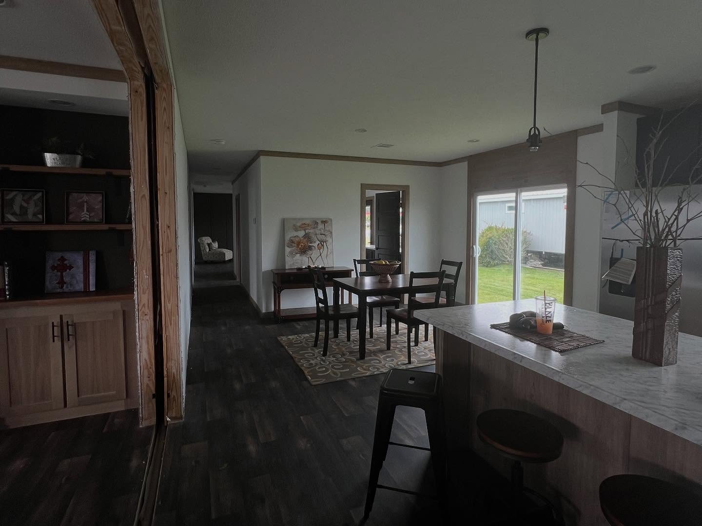 Modern dining area with a wooden table and chairs on a patterned rug, next to large glass doors showing a green lawn. The kitchen island has a marble countertop, decor, and two stools, creating a cozy, inviting atmosphere.