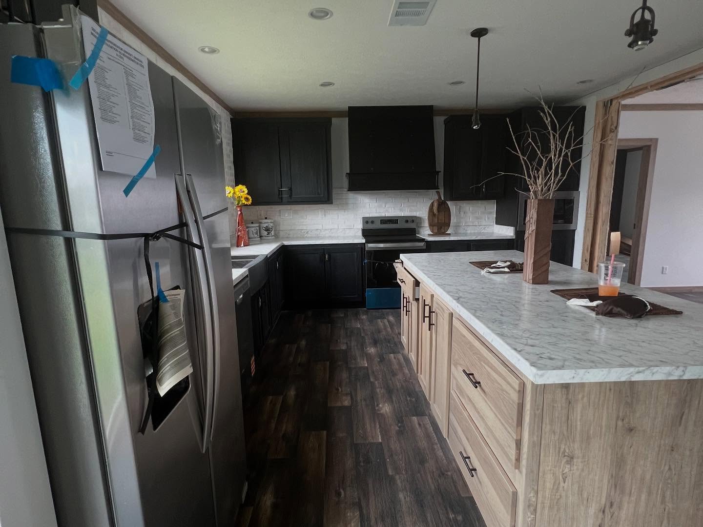 Modern kitchen with dark cabinets, marble countertops, and wooden floor. An island with decorative branches and a cup. Light and airy ambiance.