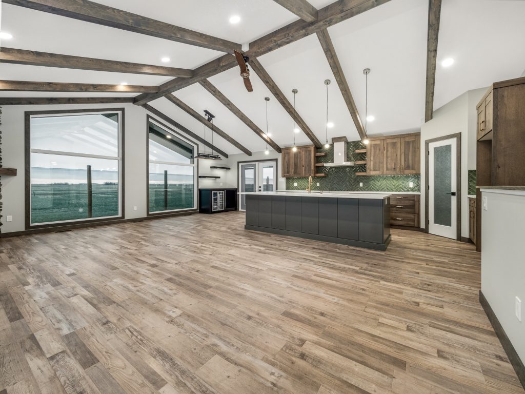 Spacious modern kitchen with wooden beams, large windows, and sleek gray cabinetry. The room features a center island and hardwood floors, evoking a warm, open feel.