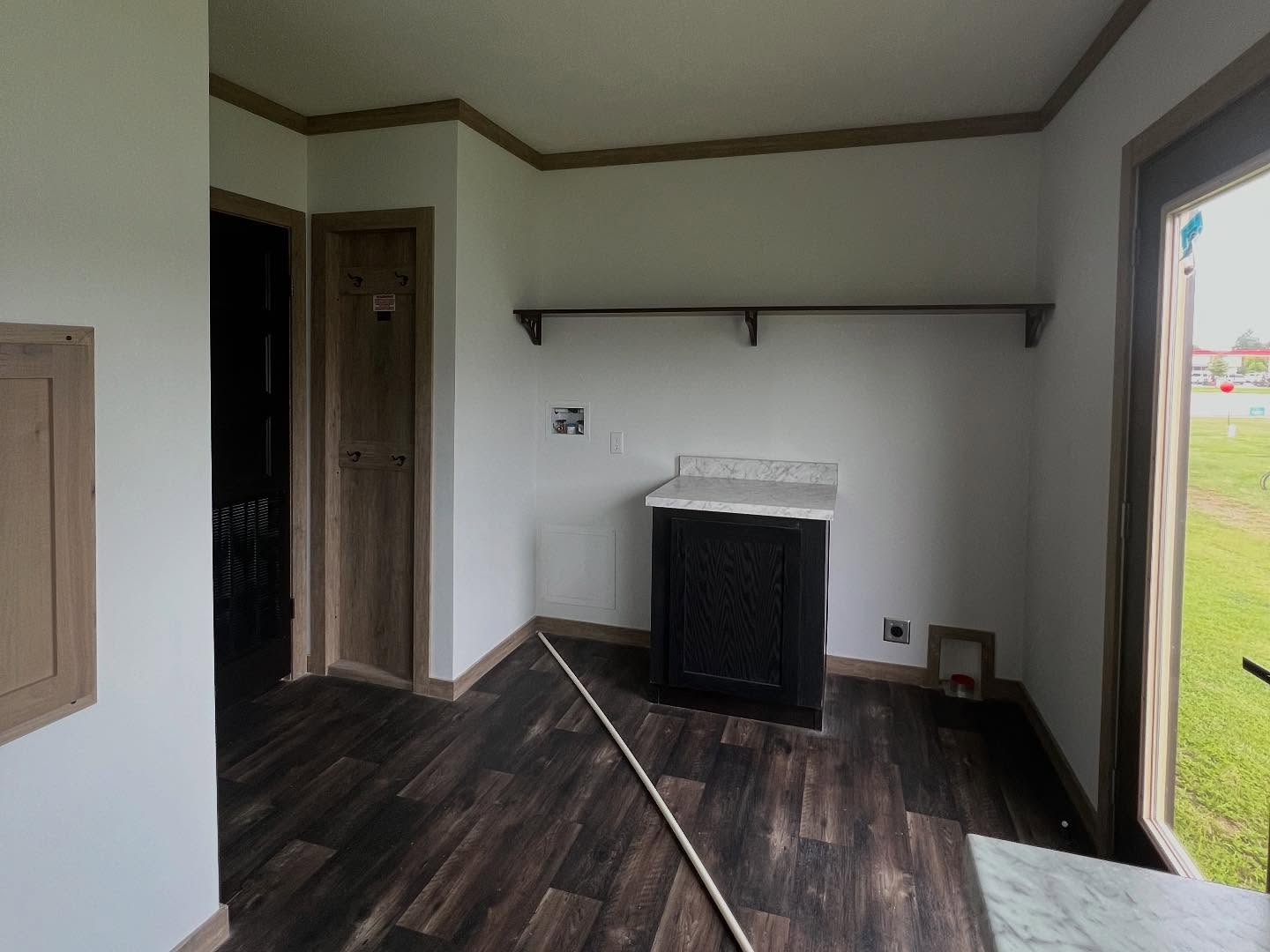 A small room with dark wood flooring and white walls. There's a black cabinet with a marble top, a wooden door, and a shelf. A large window lets in light.