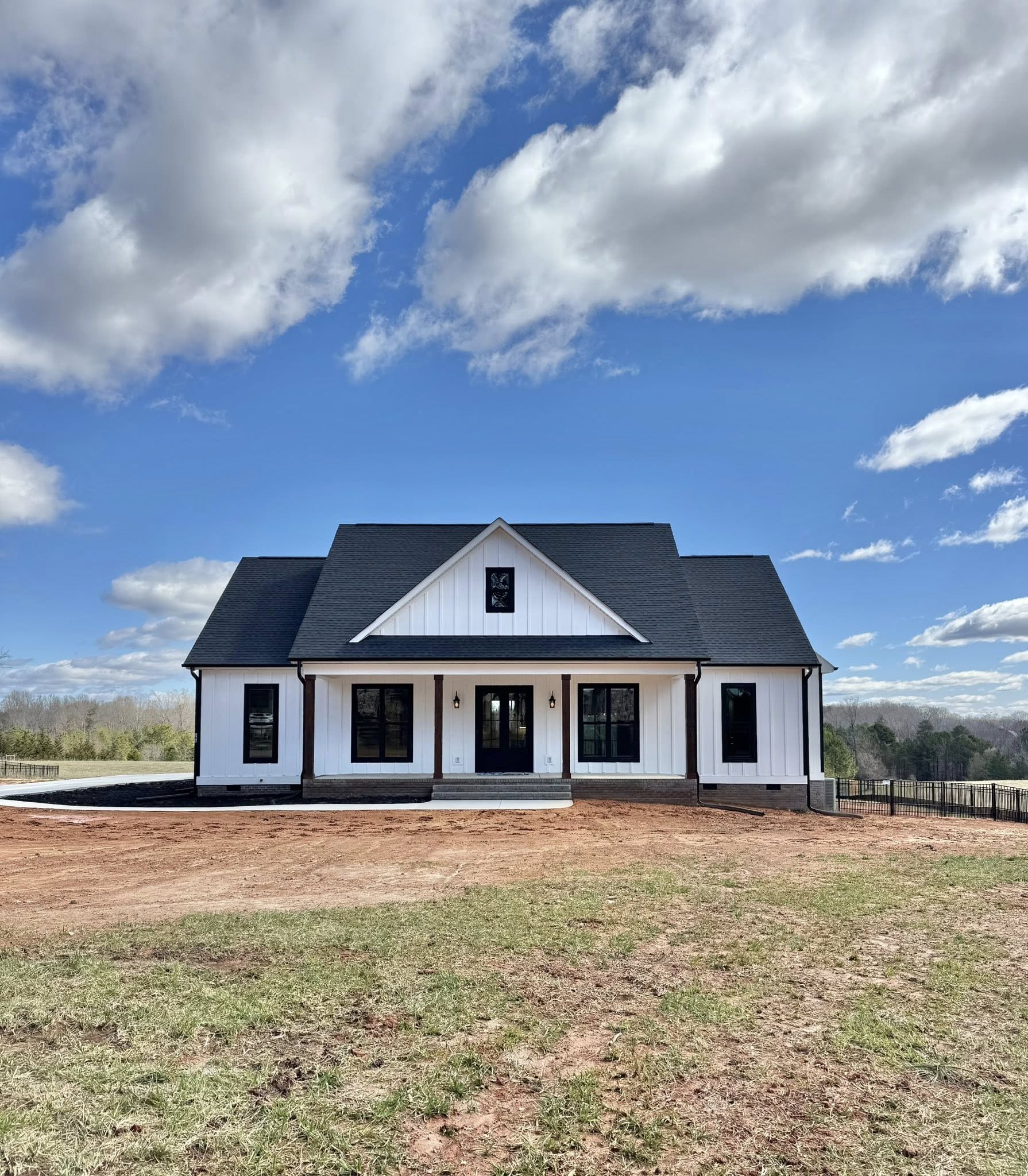 A modern farmhouse with a black roof and white siding sits under a bright blue sky with fluffy clouds. The front yard is a mix of red soil and grass.
