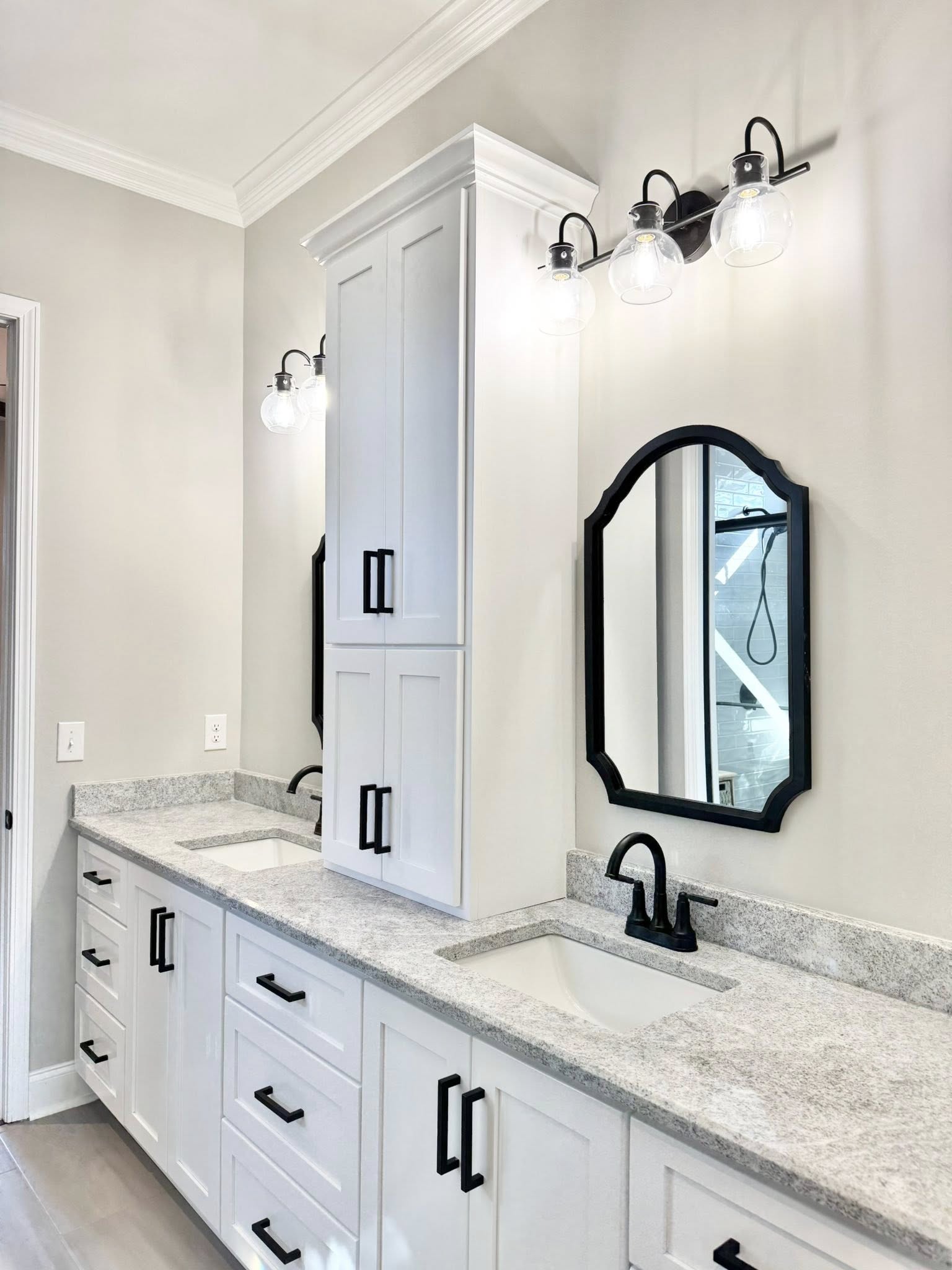 Modern bathroom with dual sinks, white cabinets, and black hardware. Oval mirrors and globe lights create a sleek, minimalist ambiance.