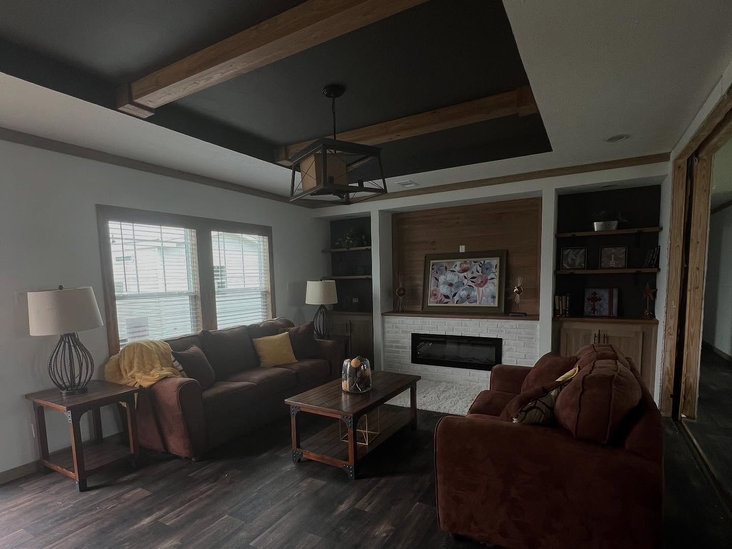 Cozy living room with dark brown sofas and a wooden coffee table. A modern ceiling light hangs above. Fireplace with floral art, shelves, and table lamps add warmth.