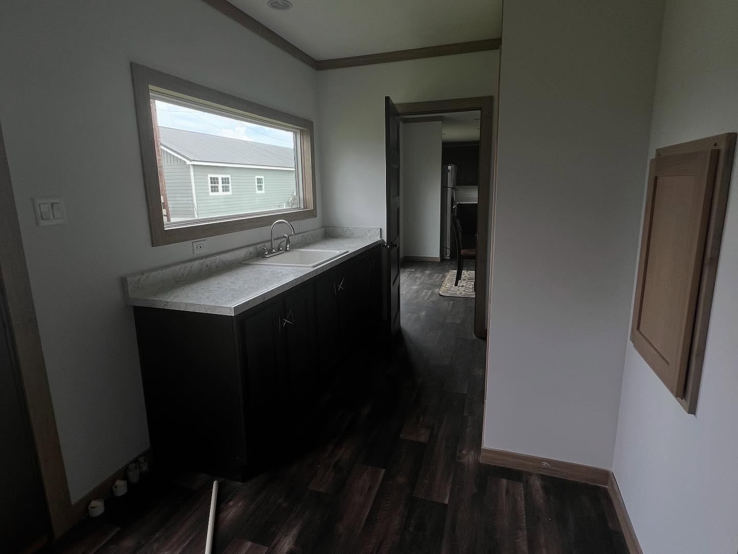 A small, dimly lit kitchen nook with dark wood floors and cabinets. A single large window illuminates a white countertop with a sink.