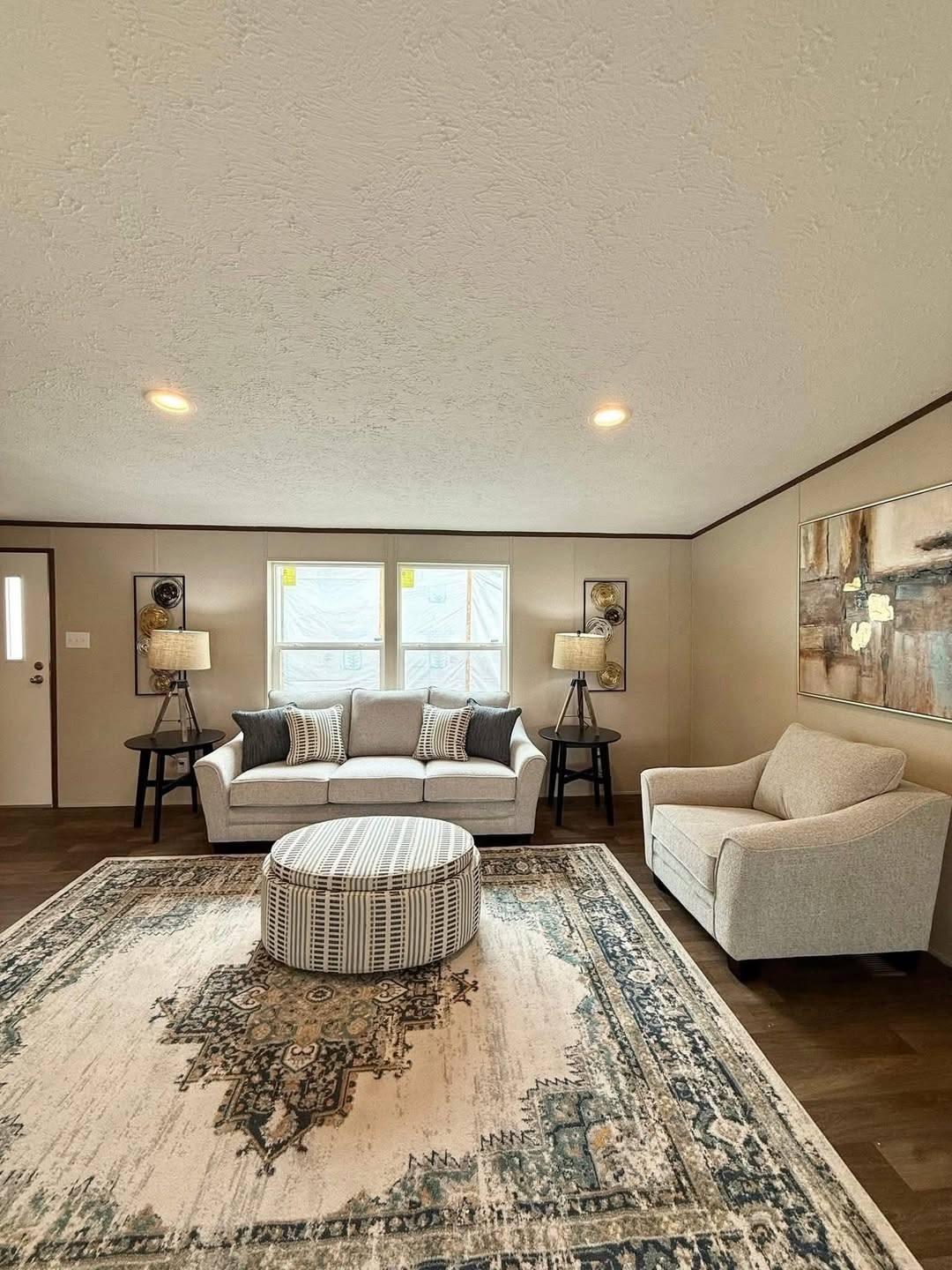 Warm living room with a gray sofa and chair facing a patterned ottoman on an ornate rug. Two lamps flank the sofa, and abstract art hangs on the wall.