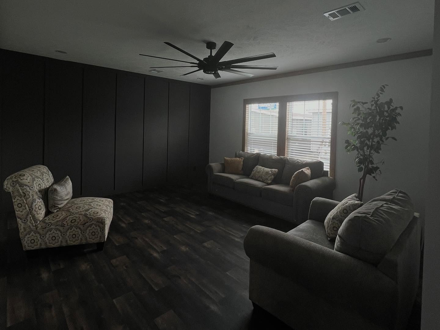 Dimly lit living room with dark wood flooring, featuring a patterned armchair, gray sofas with pillows, large ceiling fan, windows with blinds, and a potted plant.