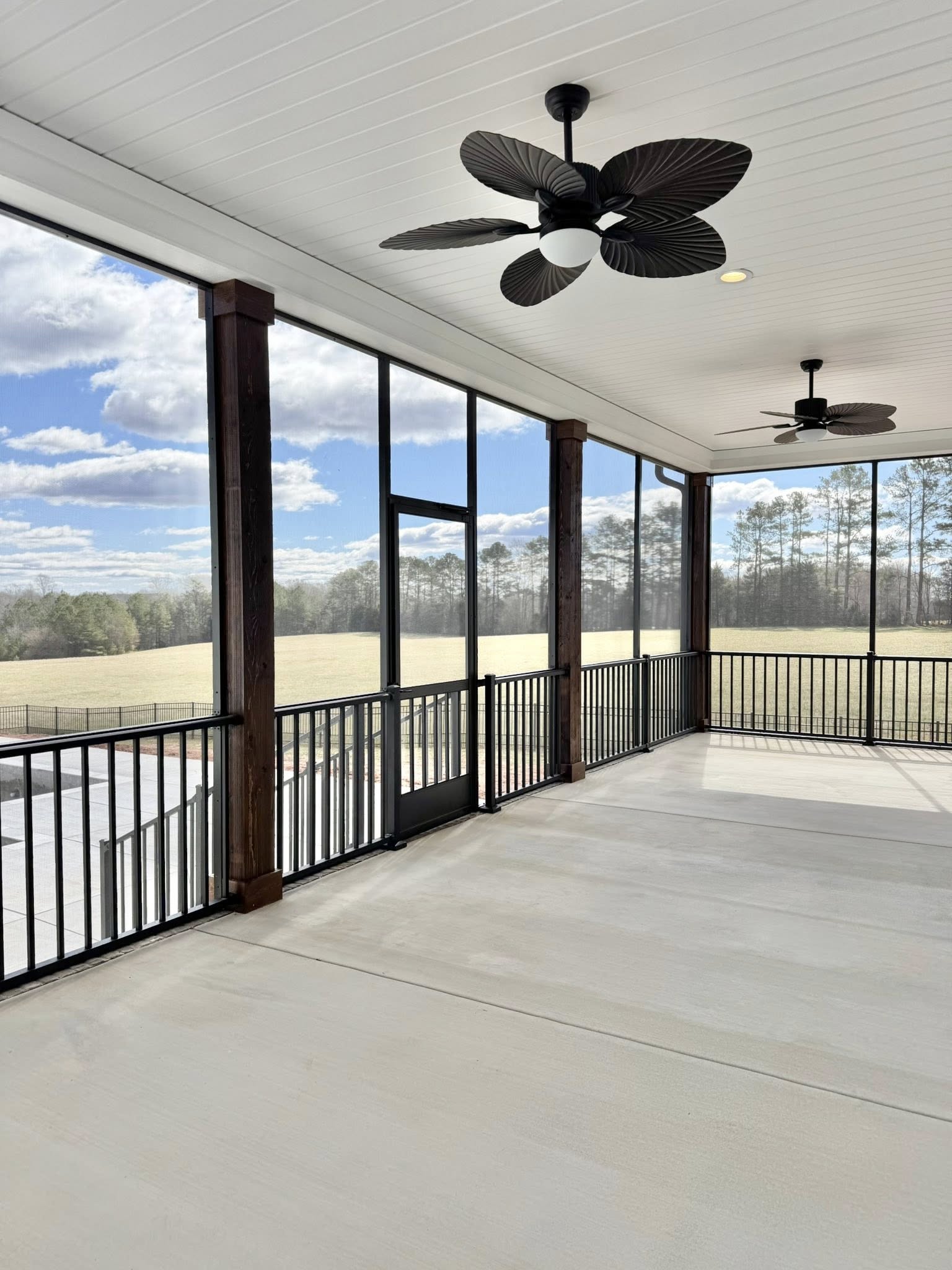Spacious screened porch with ceiling fans, overlooking a sunny field and distant trees. The bright sky is filled with scattered white clouds.