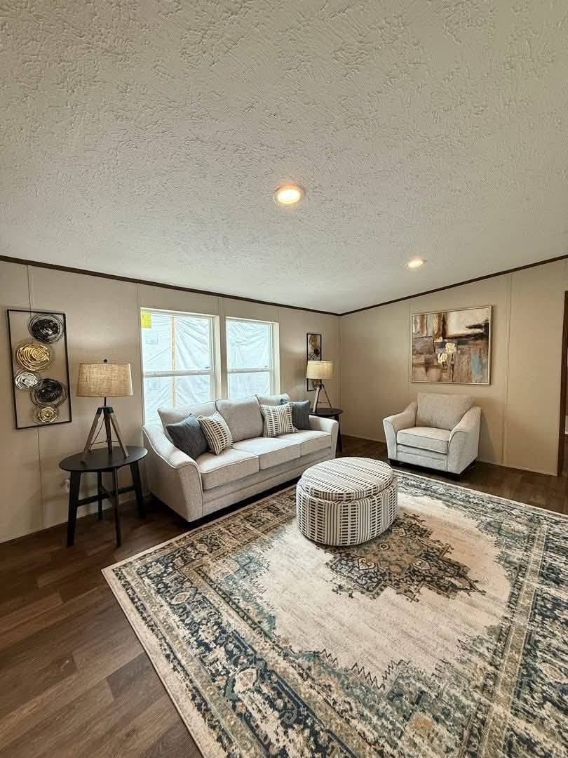 A cozy living room with neutral tones features a white sofa, armchair, and round ottoman on a patterned rug. Warm light from lamps and wall decor creates a welcoming atmosphere.