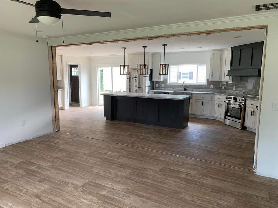 Open-plan kitchen with wooden flooring. Features a black island, pendant lights, white cabinets, and stainless steel appliances. Bright, modern, and inviting.